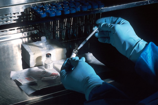 two gloved arms in view, one holding a DNA vial while the other is using a syringe to fill the vial.