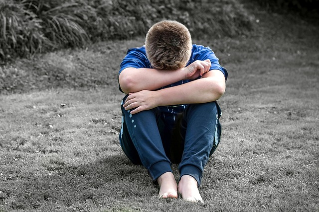 boy sitting on a lawn with his legs polled up to his chest, his head resting on his arms which are resting on his knees.