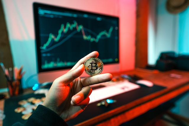 Computer in the background with what looks like a financial history diagram, and a man's hand in the foreground with a Bit Coin coin between his index finger and the middle finger.