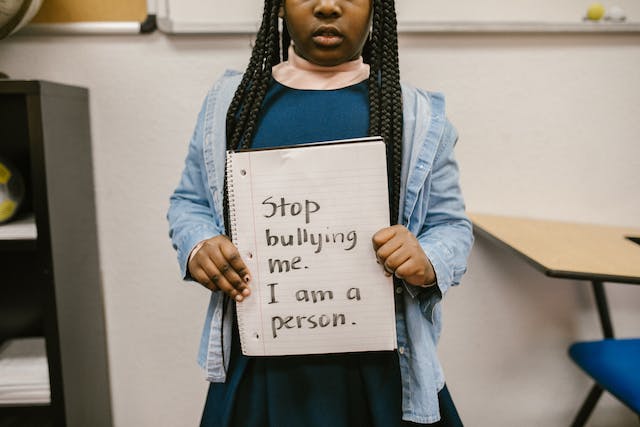 Young girl of 8 to 12 years-old standing in a classroom, it shows her from just below her eyes to her waist. She's holding a notebook with some words printed in marker. It says "Stop bullying me. I'm a person."