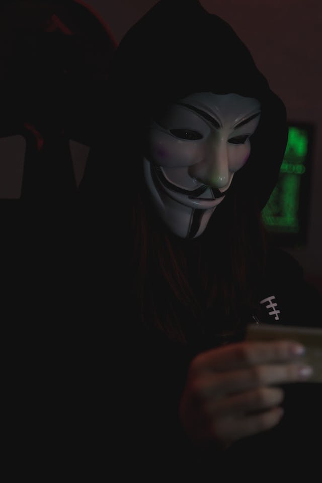 Man dressed in a hoodie with a guy fox mask on, he's holding what appears to be a card.