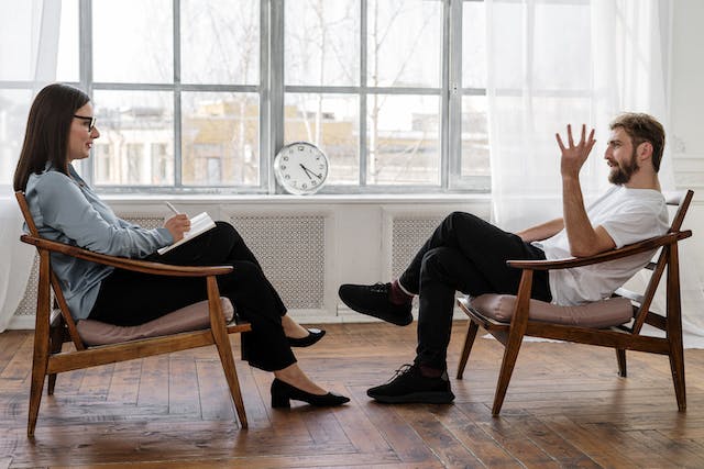 Two people sitting in a chair, the woman on the left is taking notes as the man on the right makes hand gestures. It's a therapy session.