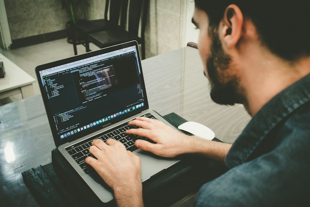 Man sitting at a table with a laptop and mouse the screen has code on it.
