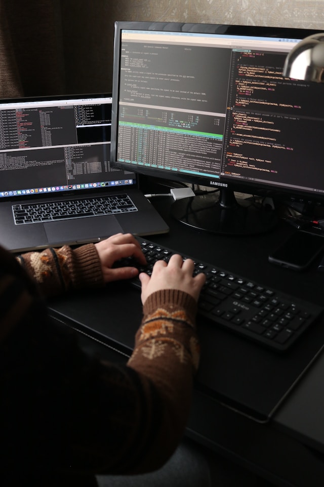 Person sitting at a desk with a laptop and a monitor and keyboard. On the screens there is windows with programming code.