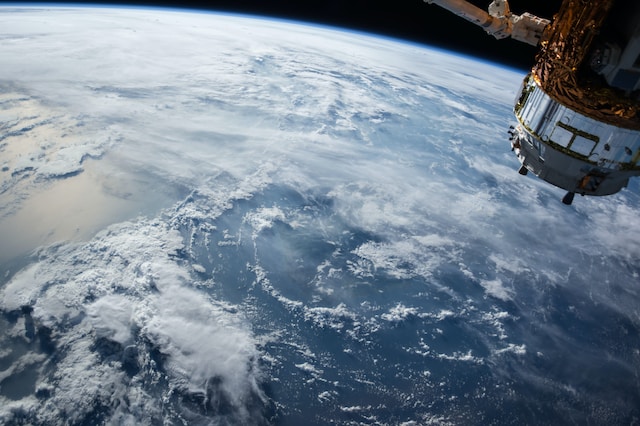 View of earth with clouds, in the top right of the image there is a satellite pointing down towards the Earth.