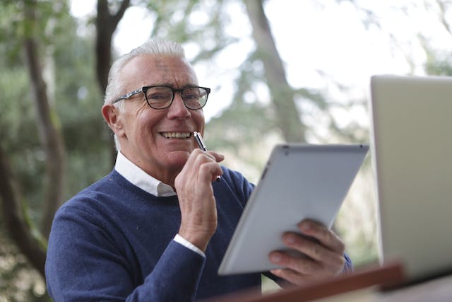 Senior man sitting holding what appears to be a Apple iPad, smiling at the camera, while holding a pen near his face.