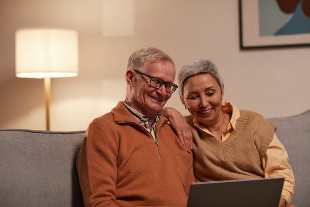 Two seasoned adults sitting on a couch, they are both smiling and looking down at the screen of a laptop.
