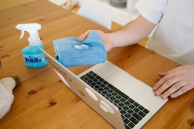 Man sitting at a table, there's a laptop and a bottle of cleaner on the table, the man is holding a rag with cleaner ready to wipe the laptop screen.