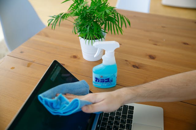 Man cleaning a laptop monitor with a towel, on the desk next to the laptop is a plant and a bottle of cleaner.