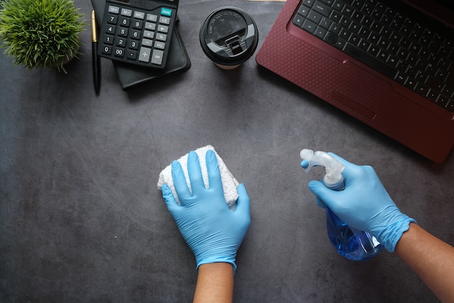 Downward view of a desk with a laptop, coffee cup, calculator, pen and a small round plant. Two hands, covered in cleaning gloves, with the left hand holding a rag and the right hand holding a bottle of cleaner. They are cleaning the desk.