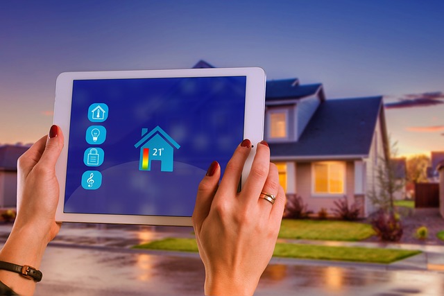 Close up of a woman's hands holding up a tablet with controls for a smart home, in the background is a house we assume is being controlled.